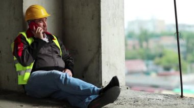 İşçi akıllı telefondan konuşur. Sarı şapkalı inşaatçı inşaat alanında yerde oturuyor ve cep telefonuyla konuşuyor.