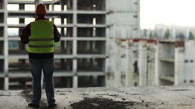 İnşaatçı yeni şantiyeye bakıyor. Başlıklı işçi proje sitesinde duruyor ve gün batımındaki yeni bina yapısına bakıyor.