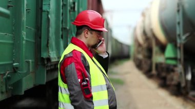 Demiryolu işçisi tren yakınlarında cep telefonuyla konuşuyor. Demiryolu çalışanı demiryolu hattında çalışıyor. Cep telefonuyla konuşuyor. İşyerinin müdürü, nakliye istasyonundaki akıllı telefon aracılığıyla iletişim kuruyor.