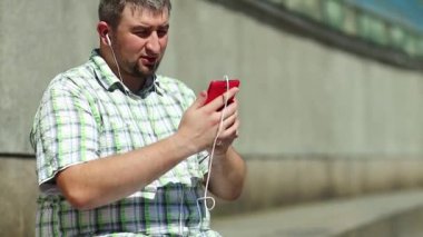 Akıllı telefonlu neşeli adam Skype üzerinden iletişim kuruyor ve el kol hareketi yapıyor. Komik adam akıllı telefondan konuşuyor. Elinde kırmızı cep telefonu olan, kareli tişörtlü, şişman, şen şakrak adam.