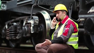 Demiryolu işçisi akıllı telefondan konuşur. Demiryolu çalışanı iş kıyafetleri içinde yük vagonunun yanında oturur ve cep telefonuyla konuşur.