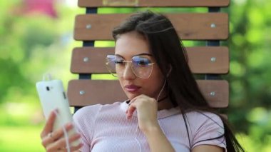 Çekici kız şehir parkında bankta oturur ve müzik dinler. Genç ve güzel bir kadın halka açık bir bahçede bankta oturur ve arka planda güzel bir güneş olan akıllı telefondan müzik dinler.