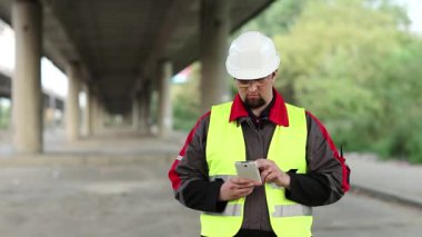 Başlıklı işçi üst geçidin altında akıllı telefon kullanıyor. İnşaat şantiyesindeki tamirat müdürünün üst yol girişleri akıllı telefonuna veri yüklüyor