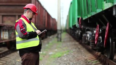 Yük trenlerinin yanında tablet bilgisayarı olan bir demiryolu işçisi. Trenler arasında tablet bilgisayarı olan bir demiryolu işçisi. Yük istasyonundaki demiryolu trafiği müfettişi tabletine notlar alır.