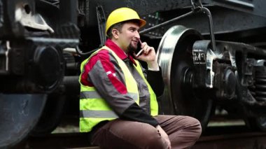 Mutlu demiryolu işçisi cep telefonuyla konuşuyor. Demiryolu işçisi yük vagonunun yanında demiryolu hattında oturur ve cep telefonuyla konuşur.