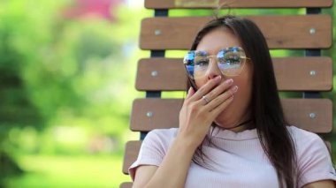 Gözlüklü güzel kadın kameraya bakar ve esner. Çekici kız şehir parkı ve esneme bankta oturur