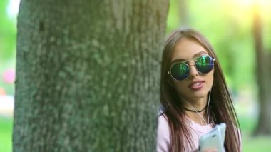 Güneş gözlüklü, kulaklıklı güzel bir kız doğada müzik dinliyor. Şehir parkında ağacın yanında duran genç bir kadın müzik dinler ve dans eder.