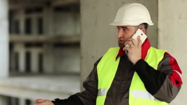 İnşaatçı inşaat alanında cep telefonuyla konuşuyor. Yeşil yelek ve beyaz şapka giyen işçiler proje sitesindeki akıllı telefondan konuşuyor