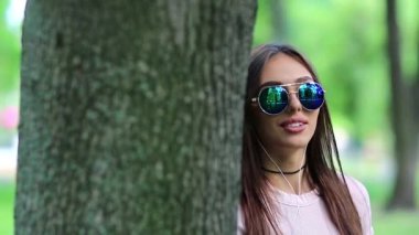 Güneş gözlüklü, kulaklıklı güzel bir kız doğada müzik dinliyor. Şehir parkında ağacın yanında duran genç bir kadın müzik dinler ve dans eder.