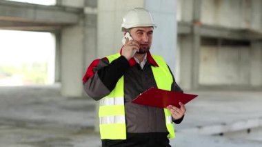 İnşaatçı inşaat alanında cep telefonuyla konuşuyor. Yeşil yelek ve beyaz şapka giyen işçiler proje sitesindeki akıllı telefondan konuşuyor