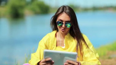 Kız gölün yanında oturuyor ve tablet bilgisayarla konuşuyor. Sarı spor ceketli sporcu kadın göletin yanında oturuyor ve tableti elinde tutuyor. Spor kıyafetli bir kadın video kaset aracılığıyla iletişim kuruyor.