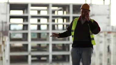 İnşaatçı inşaat alanında cep telefonuyla konuşuyor. Yeşil yelek ve beyaz şapka giyen işçiler proje sitesindeki akıllı telefondan konuşuyor