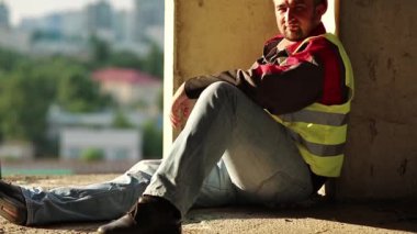 İnşaatçı yere oturur ve kameraya bakar. Sarı başlıklı müteahhit proje alanındaki beton zeminde oturuyor. İşçi inşaat alanında dinleniyor