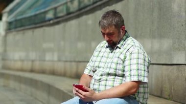 Adam akıllı telefon kullanıyor ve müzik dinliyor. İşadamı basamakta oturur ve akıllı telefonuyla müzik dinler. Kırmızı cep telefonlu şişman adam.