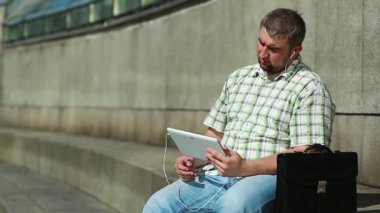 İşadamı tablet bilgisayarla iletişim kuruyor. Tabletli adam video kaset aracılığıyla iletişim kuruyor. Evrak çantalı, kareli gömlekli şişman bir adam elinde tabletle kulaklık tutuyor.