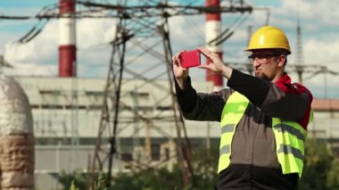 İşçi akıllı telefondan video kaydediyor. Elektrik santralinde çalışan, sarı şapkalı, akıllı telefonlu bir işçi.