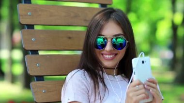 Şehir parkında bankta oturan güzel bir kadın müzik dinler, gülümser ve dans eder. Genç güzel kız halka açık bir bahçede bankta oturur ve akıllı telefondan müzik dinler.