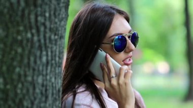 Güzel bir kadın cep telefonuyla konuşur. Uzun saçlı çekici kız ağacın yanında duruyor ve cep telefonuyla konuşuyor. Doğada akıllı telefonu olan mavi güneş gözlüklü bir iş kadını.