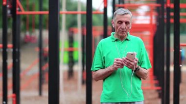 Cep telefonu olan son sınıf öğrencisi spor sahasının yanında duruyor. Aktif yaşlı sporcu cep telefonunu tutuyor. Yaşlanmış sporcu akıllı telefondan müzik dinler. Açık hava spor salonundaki gri saçlı adam.