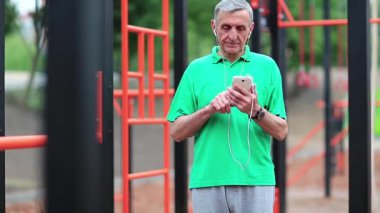 Yaşlı sporcu spor alanında akıllı telefon kullanır. Kıdemli adam cep telefonuyla çalışıyor. Gri saçlı aktif atlet açık hava spor salonunun yanında duruyor ve cep telefonunu tutuyor.