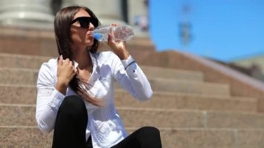 Kız merdivenlerde oturur ve şişeden su içer. Güneş gözlüklü uzun saçlı güzel kız temiz su içiyor. Kız plastik su şişesini açar ve soda içer.