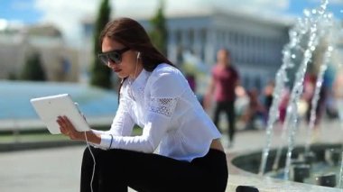 Kadın çeşmelerin yanında oturuyor ve tablet bilgisayar kullanıyor. Elinde beyaz tablet bilgisayarla gözlüklü çekici bir kadın.