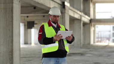 Builder speaks via tablet pc on project site. Worker with tablet computer on construction site. Workman in white hard hat holds in hand personal computer, looks in display