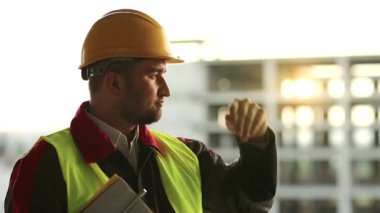 Proje alanında inşaat belgelerine sahip bir inşaatçı. İnşaat alanındaki inşaat belgeleriyle birlikte kasketli usta inşaatçı ileriye bakıyor.