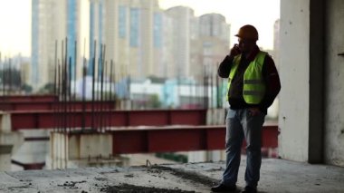 Foreman inşaat alanında cep telefonuyla konuşuyor. Yeşil yelek giyen inşaatçı proje sitesinde telefonla konuşuyor