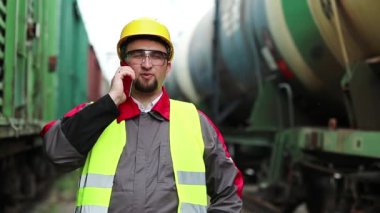 Demiryolu işçisi tren yakınlarında cep telefonuyla konuşuyor. Demiryolu çalışanı demiryolu hattında çalışıyor. Cep telefonuyla konuşuyor. İşyerinin müdürü, nakliye istasyonundaki akıllı telefon aracılığıyla iletişim kuruyor.