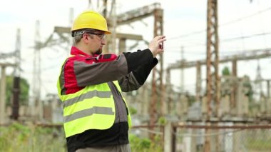 Elektrikçi akıllı telefondan video kaydediyor. Elektrik santralinde akıllı telefonlu sarı şapkalı güçlü adam.