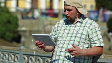 Adam tablet bilgisayarla iletişim kuruyor. Kareli gömlekli ve beysbol şapkalı şişman çocuk elinde kulaklıkla beyaz tablet tablet tutuyor. Tabletli adam görüntülü sohbet yoluyla iletişim kuruyor