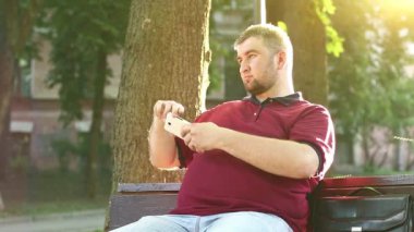 İş adamı bankta oturur ve akıllı telefondaki videoları kaydeder. Şişman adam cep telefonuyla fotoğraf çekiyor.