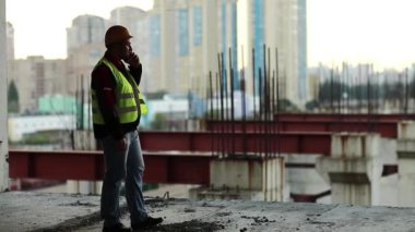 Foreman inşaat alanında cep telefonuyla konuşuyor. Yeşil yelek giyen inşaatçı proje sitesinde telefonla konuşuyor