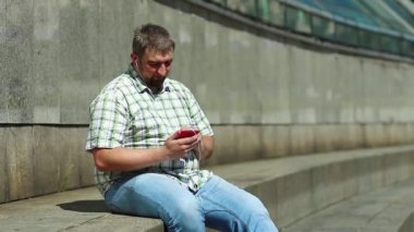 Adam akıllı telefon kullanıyor ve müzik dinliyor. İşadamı basamakta oturur ve akıllı telefonuyla müzik dinler. Kırmızı cep telefonlu şişman adam.