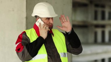 İnşaatçı inşaat alanında cep telefonuyla konuşuyor. Yeşil yelek ve beyaz şapka giyen işçiler proje sitesindeki akıllı telefondan konuşuyor