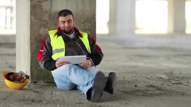 Tablet bilgisayarı olan işçi inşaat alanında oturur. İnşaatçı proje sitesindeki tablet bilgisayarla konuşuyor. Beyaz başlıklı işçi elinde kişisel bilgisayarını tutuyor, görüntüye bakıyor ve konuşuyor.