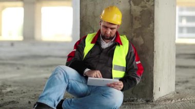 Tablet bilgisayarı olan inşaatçı yere oturur. İnşaat alanında tablet bilgisayarla çalışan işçiler. Beyaz şapkalı mimar beton zeminde oturur ve elinde kişisel bilgisayarla ekranda görünür.