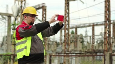 İşçi akıllı telefondan video kaydediyor. Elektrik santralinde akıllı telefonlu sarı şapkalı güçlü adam.