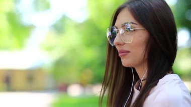 Akıllı telefonu ve kulaklığı olan gözlüklü güzel bir kadın müzik dinliyor. Akıllı telefonlu kız şehir parkında müzik dinliyor.