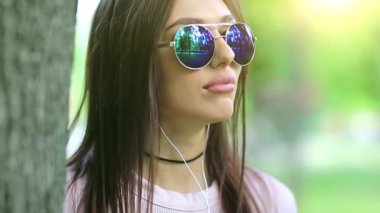 Güneş gözlüklü, kulaklıklı güzel bir kız doğada müzik dinliyor. Şehir parkında ağacın yanında duran genç bir kadın müzik ve dans dinliyor, arka planda güzel bir güneş var.