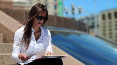 Ticari evrakları olan konsantre bir kadın. Genç kızgın kariyer kadını şehir merkezindeki büyük merdivenlerde oturuyor ve elinde iş belgeleriyle geziyor, rüzgar ona esiyor, stresli.