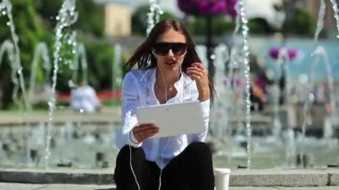 İş kadını çeşmelerin yanında oturur ve tablet bilgisayarla iletişim kurar. Beyaz tabletli gözlüklü çekici bir kadın Skype ya da görüntülü sohbet yoluyla iletişim kuruyor.