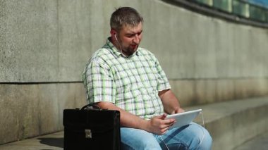 İşadamı tablet bilgisayarla iletişim kuruyor. Tabletli adam video kaset aracılığıyla iletişim kuruyor. Evrak çantalı, kareli gömlekli şişman bir adam elinde tabletle kulaklık tutuyor.