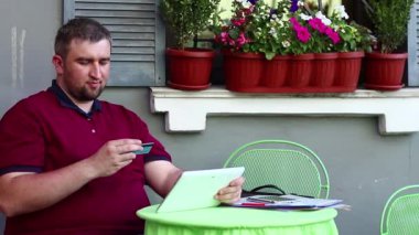 İş adamı yaz restoranında masada oturuyor, elinde tablet bilgisayar ve mavi kredi kartı tutuyor, veri giriyor. Adam kredi kartıyla online alışveriş yapıyor, kredi kartıyla alışveriş yapıyor.