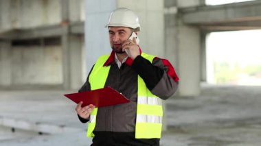 Proje sitesindeki inşaatçı cep telefonuyla konuşuyor. Beyaz başlıklı işçi inşaat alanında akıllı telefondan konuşuyor.