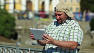 Adam tablet bilgisayarla iletişim kuruyor. Kareli gömlekli ve beysbol şapkalı şişman çocuk elinde kulaklıkla beyaz tablet tablet tutuyor. Tabletli adam görüntülü sohbet yoluyla iletişim kuruyor