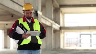 İnşaatçı proje sitesinde tablet bilgisayarla çalışıyor. İnşaat alanında tablet bilgisayarı olan bir işçi. Beyaz başlıklı işçi elinde kişisel bilgisayarla, ekranda görünüyor.