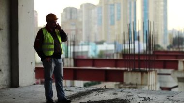 İşçi inşaat alanında akıllı telefondan konuşur. Yeşil yelek giyen inşaatçı proje alanında telefonla konuşuyor. İşçi el çantası ve cep telefonunu tutuyor.