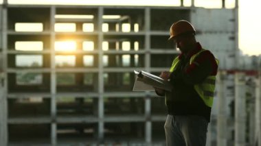 Proje sahasında çalışma belgelerine sahip bir inşaatçı. Usta müteahhit iş belgelerine notlar alır. Bina müfettişi belgelerine arka planda güzel bir güneş olduğunu yazmış.